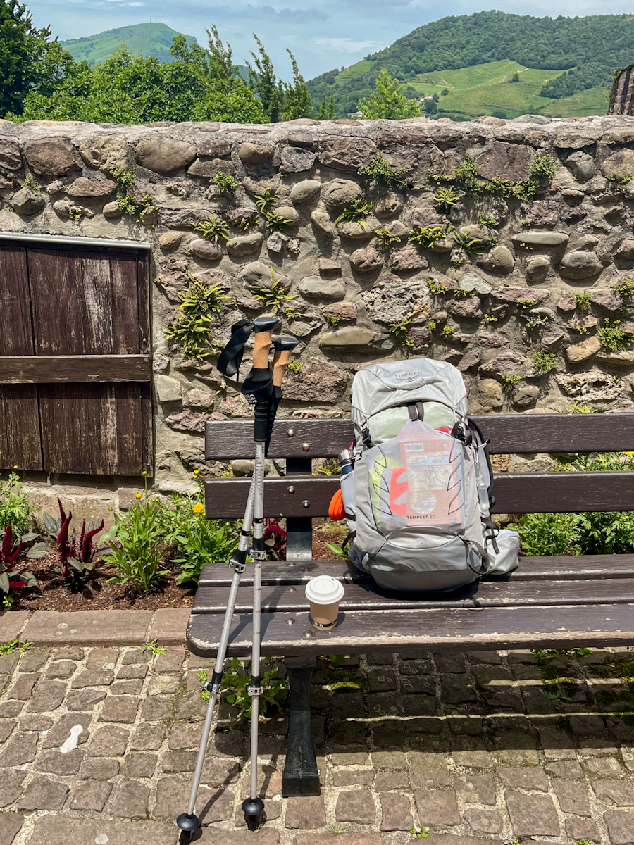 A backpack and trekking poles resting against a bench by a stone wall, a coffee cup beside it