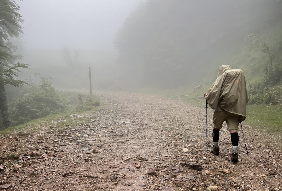 A hooded hiker in a rain poncho walking away down a stony path with trekking poles, fog ahead