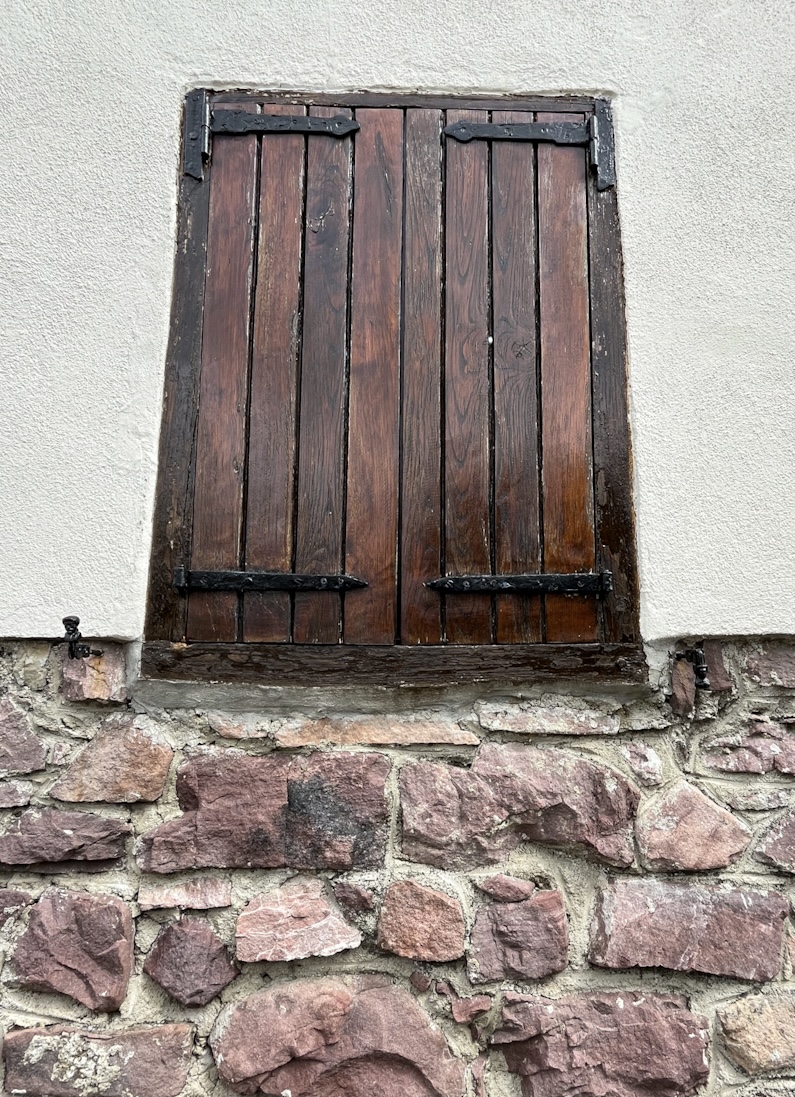 Closed wooden shutters with black iron hinges set into a whitewashed and stone wall
